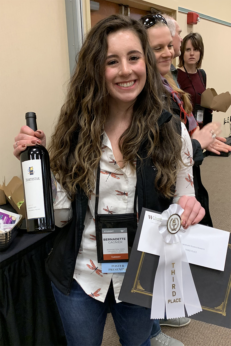Smiling young woman holds a bottle of wine and a 3rd place ribbon.