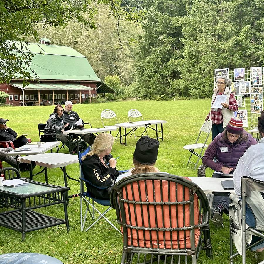 Standing person with posters and handouts speaks to seated people in an outoor, forested setting.