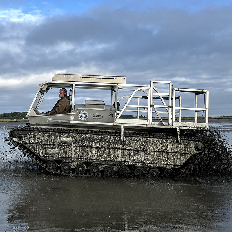 A boat-like vehicle with tank-type treads churning up sediment in shallow water.