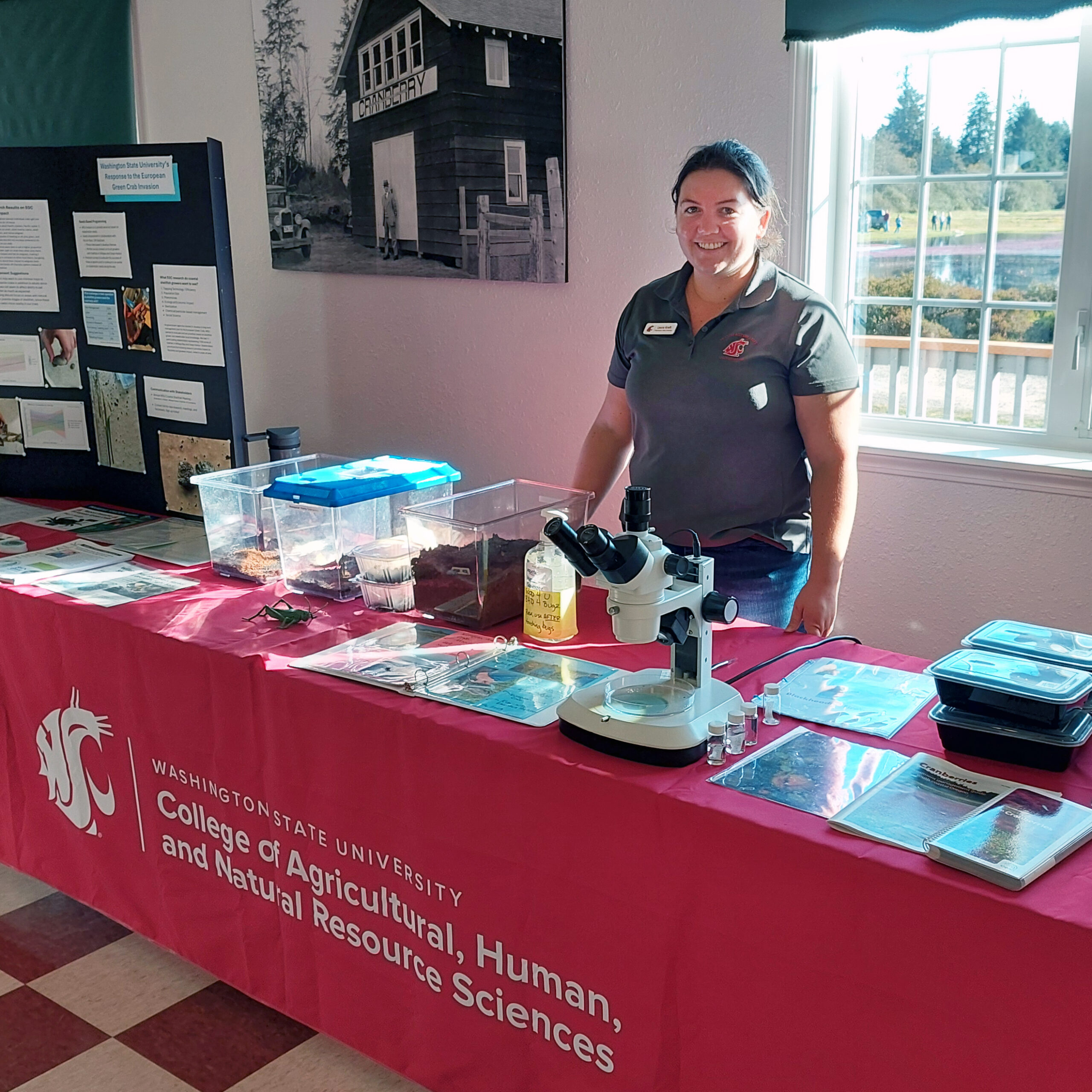 Smiling person behind a table topped with brochures, posters, and a microscope.