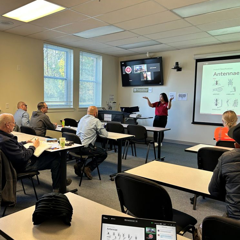 Person gesturing in front of a classroom, with slide show and Zoom screens behind them.
