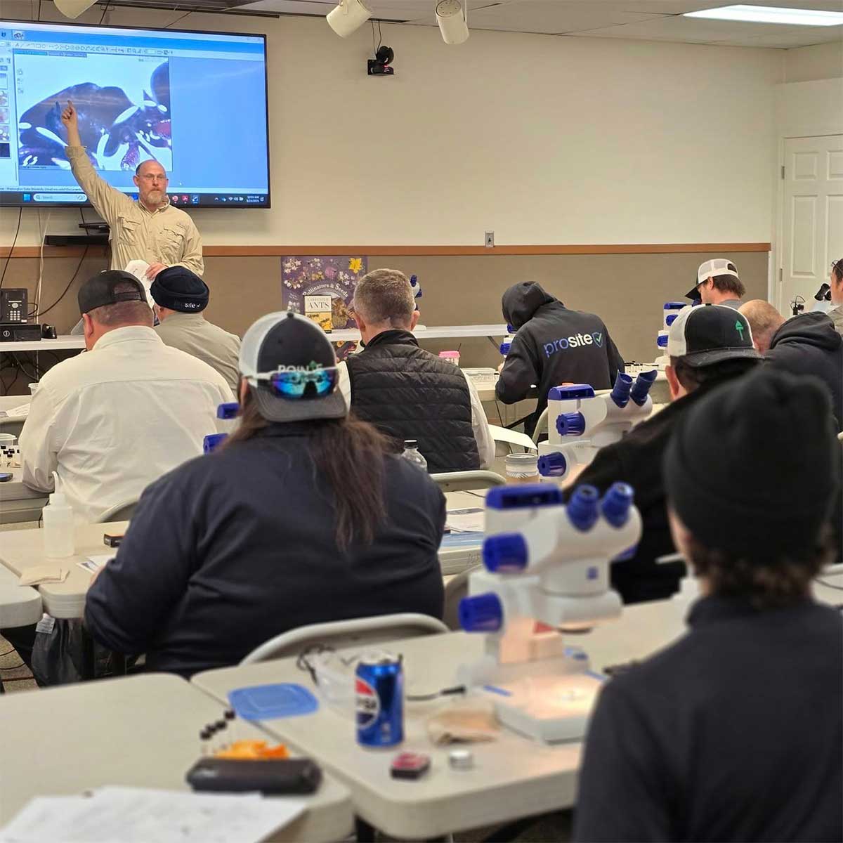 Speaker with slide show addressing a classroom of adult students with microscopes.