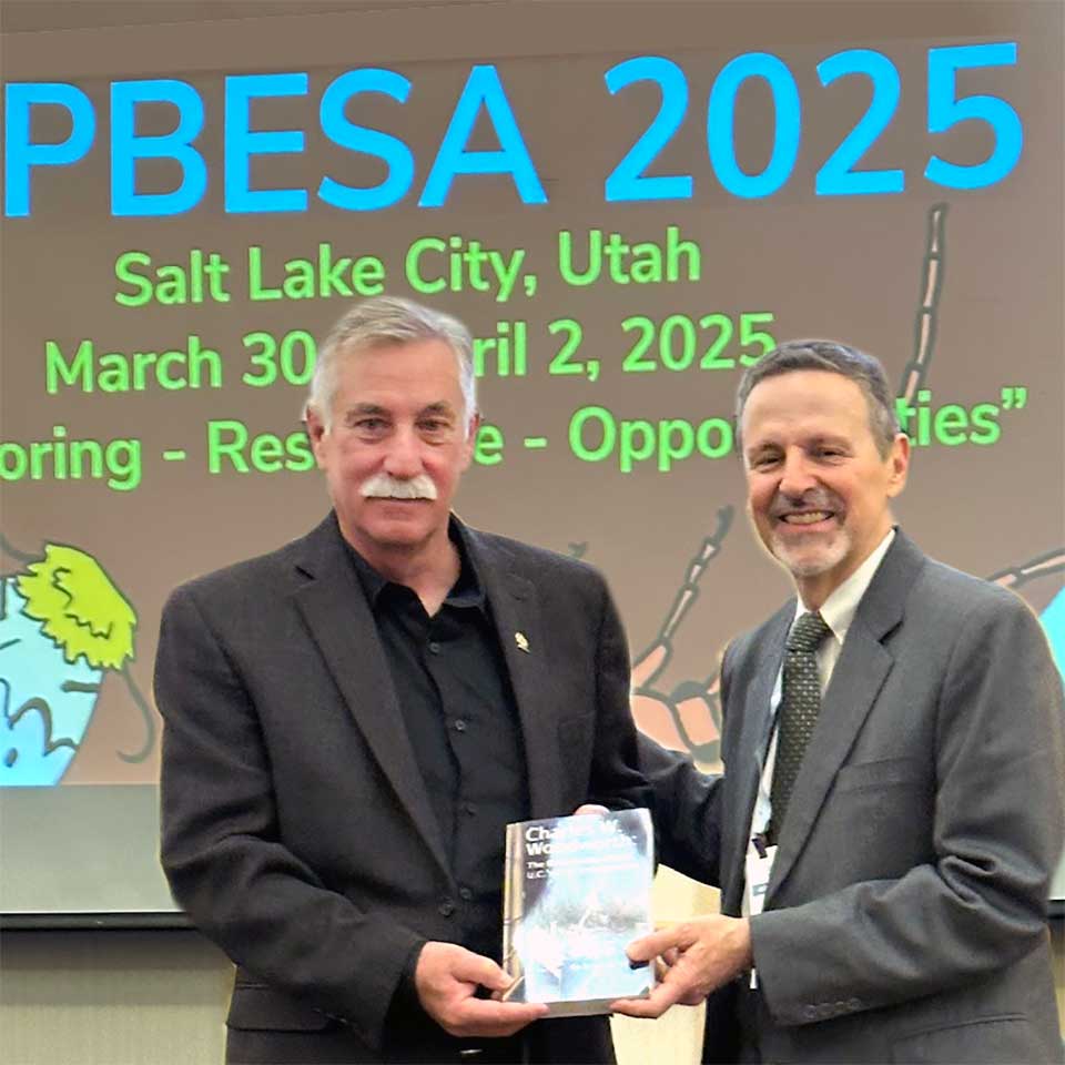 Two men holding a paperback book standing in front of a poster.