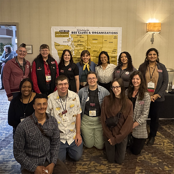 Thirteen smiling people gathered in front of a poster about bee clubs in the state of Washington.