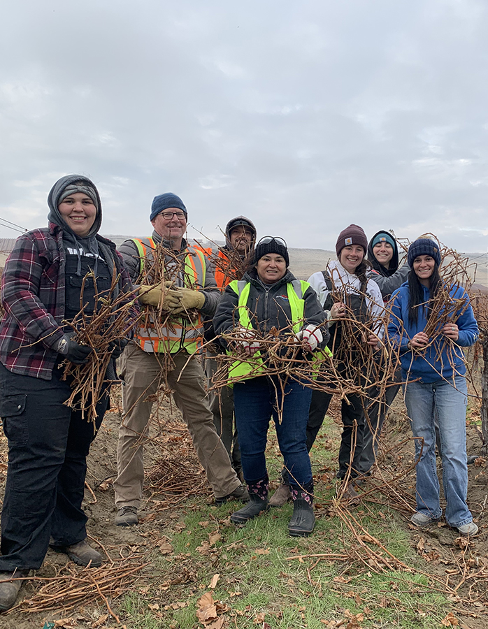 Seven smiling people in winter work clothes holding grapevine trimmings.