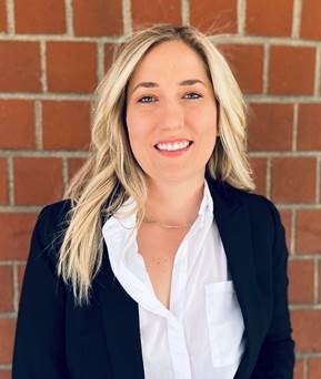Catherine J Headshot.  Catherine is standing in front of a red brick building wearing a business suit.