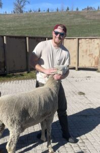Jake McNeill wearing a hat and sunglasses is standing on a brick patio with a green hill in the background holding onto the head of an affectionate sheep.
