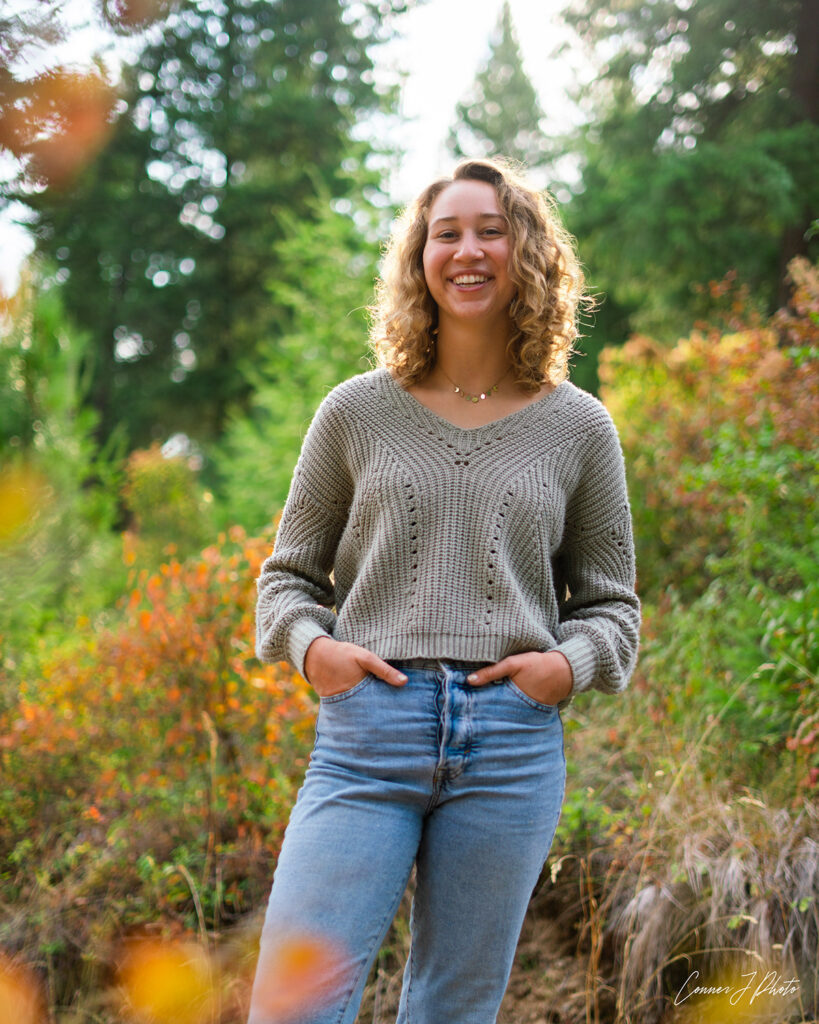 A smiling young person with light curly hair is wearing a grey sweater and jeans with her hands in her front pockets. She is standing amidst green shrubbery with trees behind her.