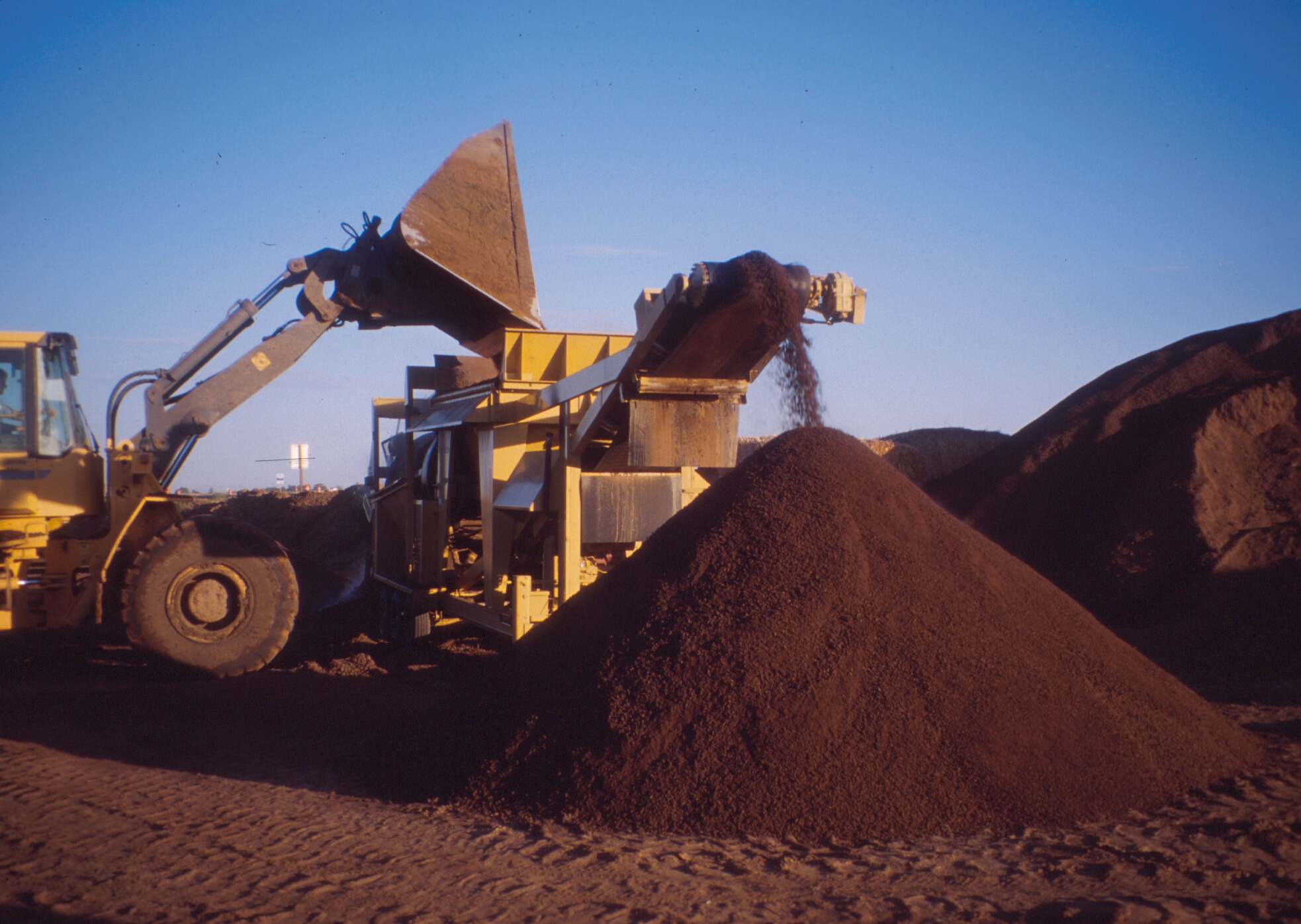 A front end loader on the left side of the screen is dumping compost into a screening machine. Screened compost is rolling off the front of the machine into a large triangular pile of screened compost.