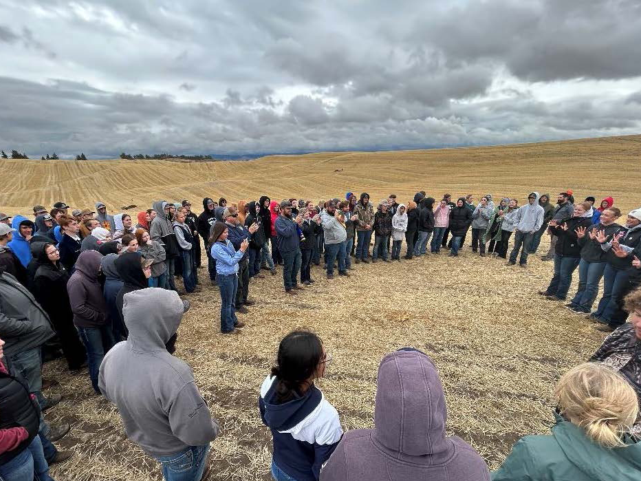 Dozens of students gather on a field under a cloudy sky.