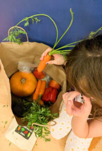 Top view of a student putting a carrot in a paper bag. there are already peppers, a small pumpkin, apples and other carrots in the bag.