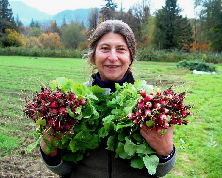 Anne Schwartz holding bunches of radishes in field with trees and mountains in the background.