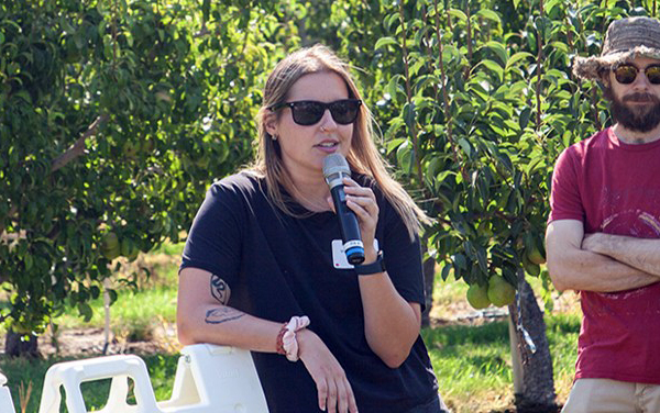 A woman with a microphone conducts an outreach session in a sunny orchard.