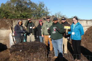 A group of seven individuals stands behind a compost pile on a sunny day.