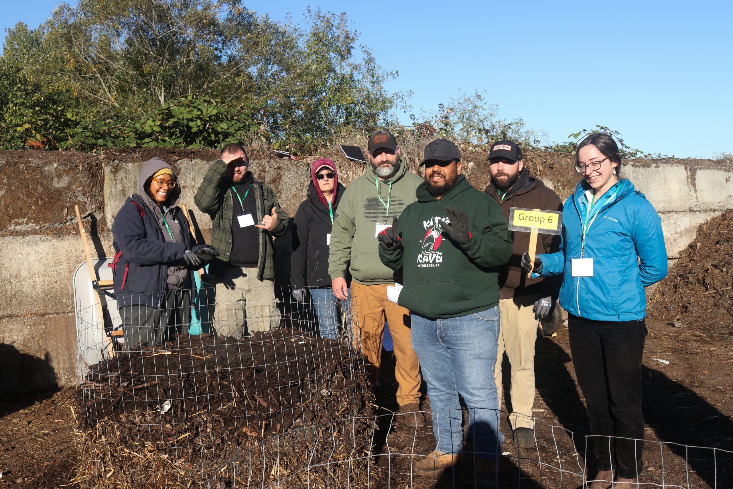A group of seven individuals stands behind a compost heap on a sunny day.