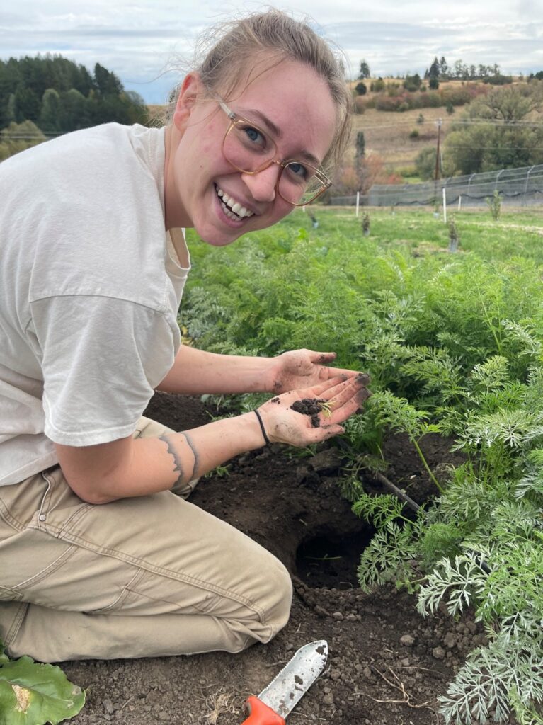 A young woman wearing glasses kneels in a field and shows the camera dirt in her hands.
