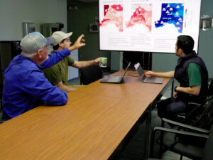 Three people look together at a screen with maps, while one points at the maps.