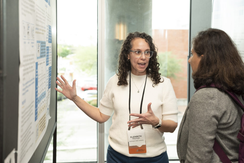One person gesturing and speaking to another person in front of a poster on the wall.