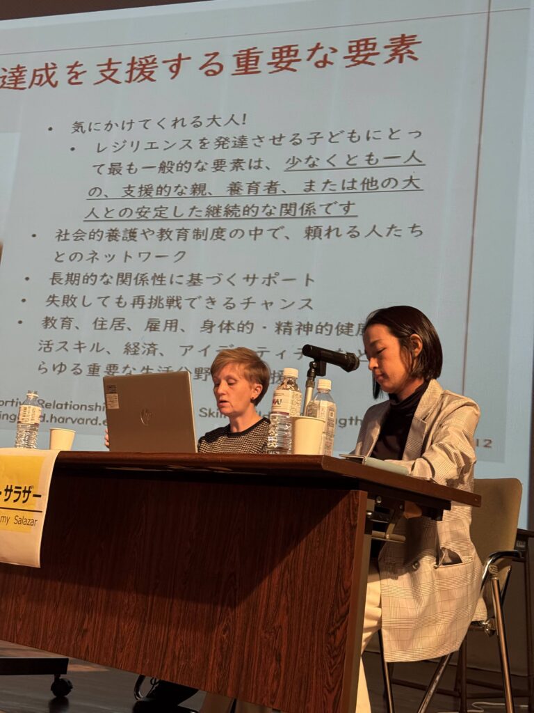 Two people sitting at a desk on a stage with a presentation screen behind them.
