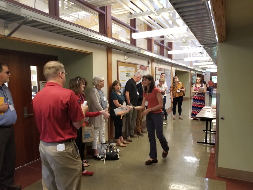 Woman speaks to group standing along wall in hallway.