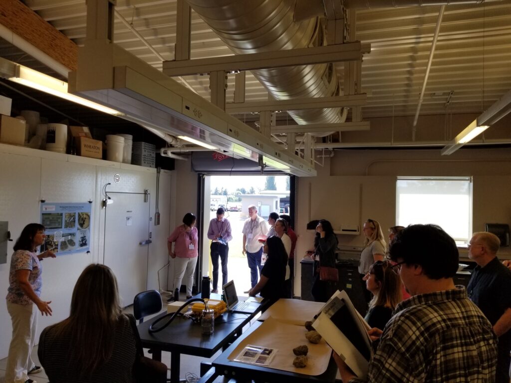 Woman speaks with group in large lab space; roll up door open to outside.