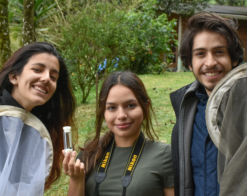 Three students from Guatemala stand and smile for a picture. One holds up a tube and the other two hold up their bug nets.