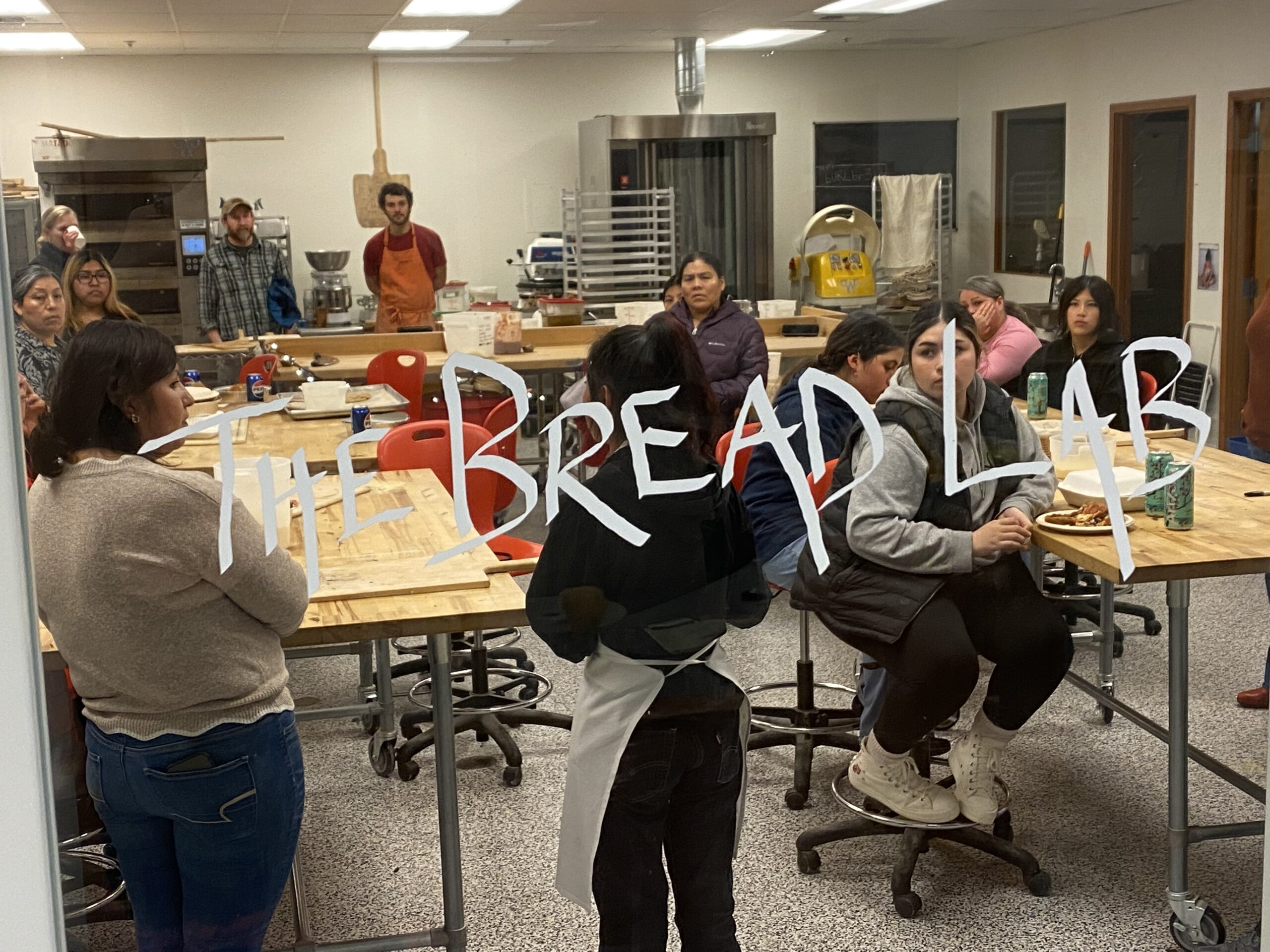 group of people listening through a window that has "the bread lab" written on it.