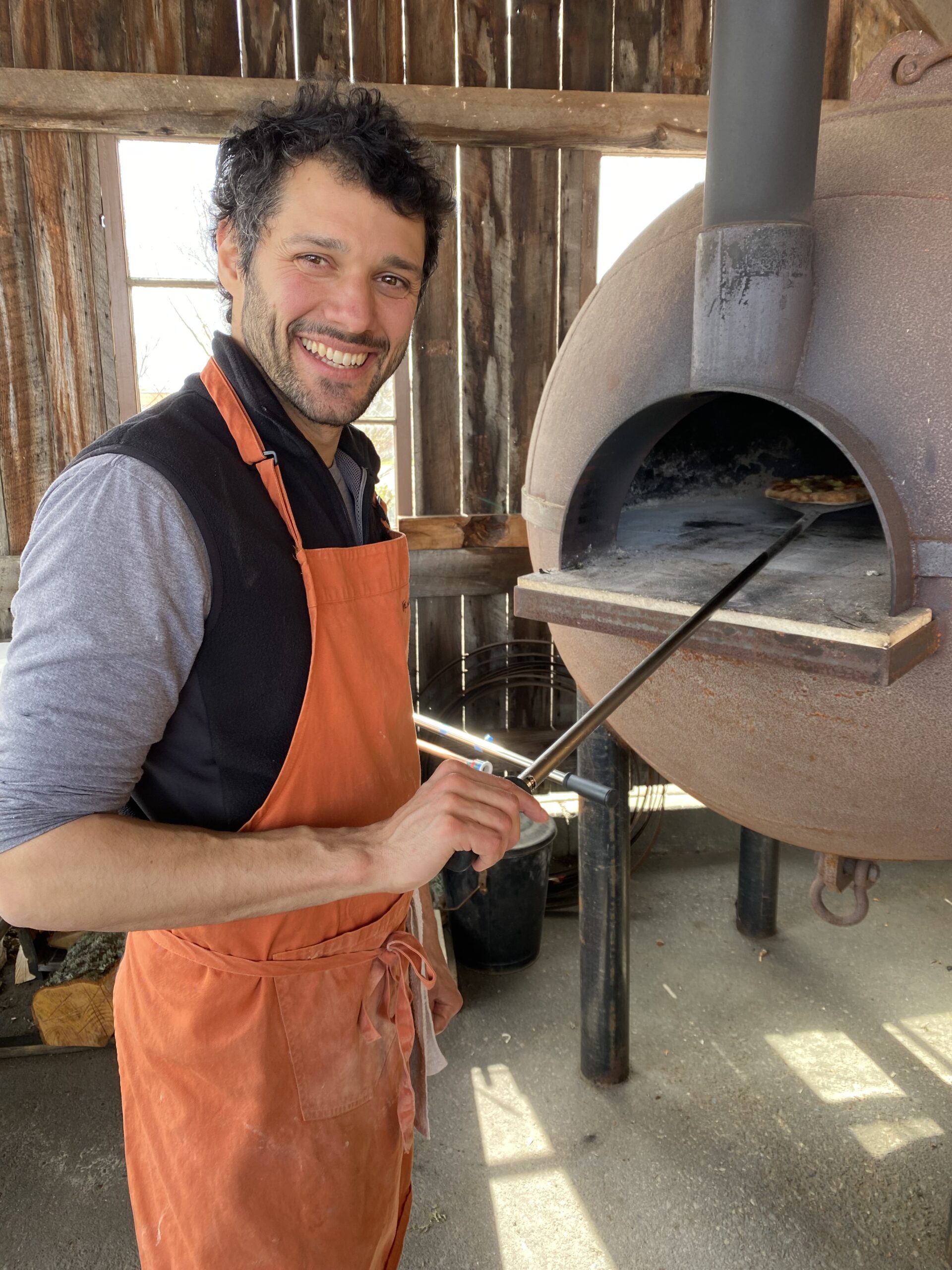 Man in an orange apron standing in front of a wood fired oven