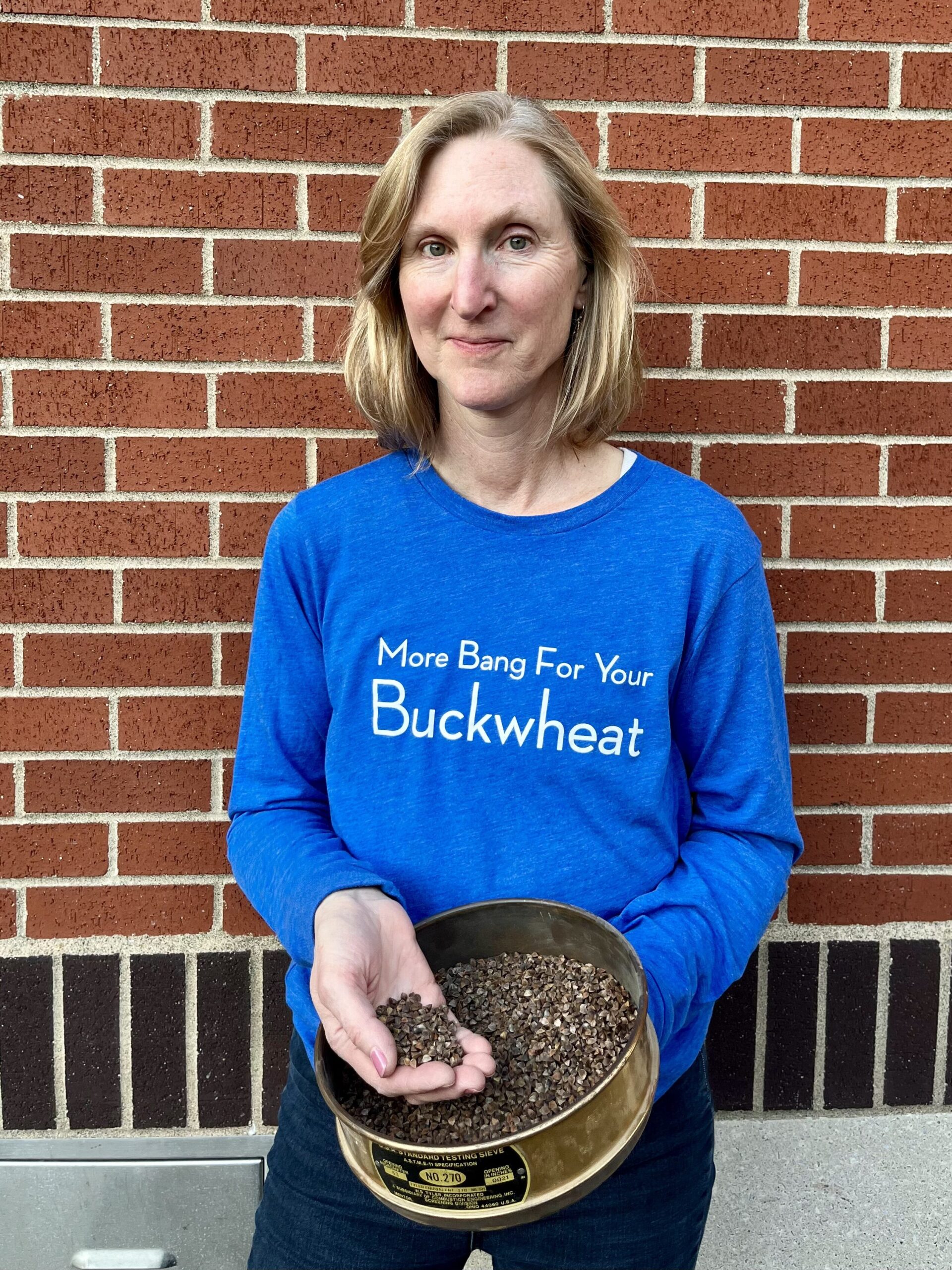 Woman in a t-shirt that reads "more bang for your buckwheat" standing against a brick wall holding a handful of grain.