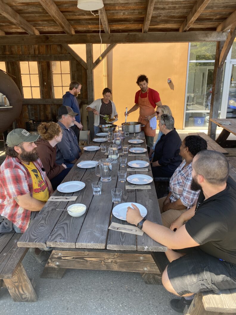 Group of 10 people sitting around a picnic table with place settings.