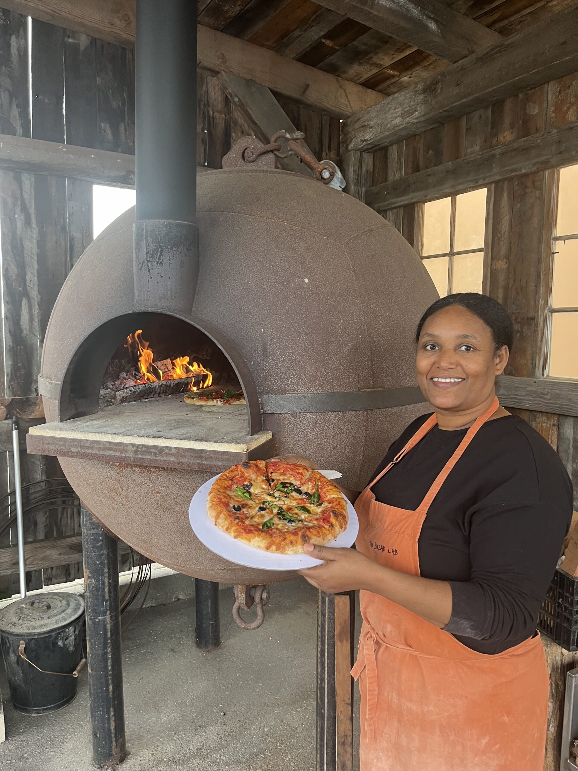 Woman stands in front of a wood-fired oven holding a cooked pizza.
