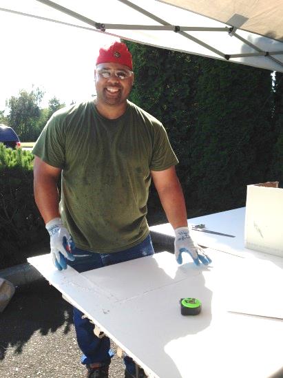 Construction laborer in safety glasses and work gloves, smiling at camera in front of measured and cut drywall.
