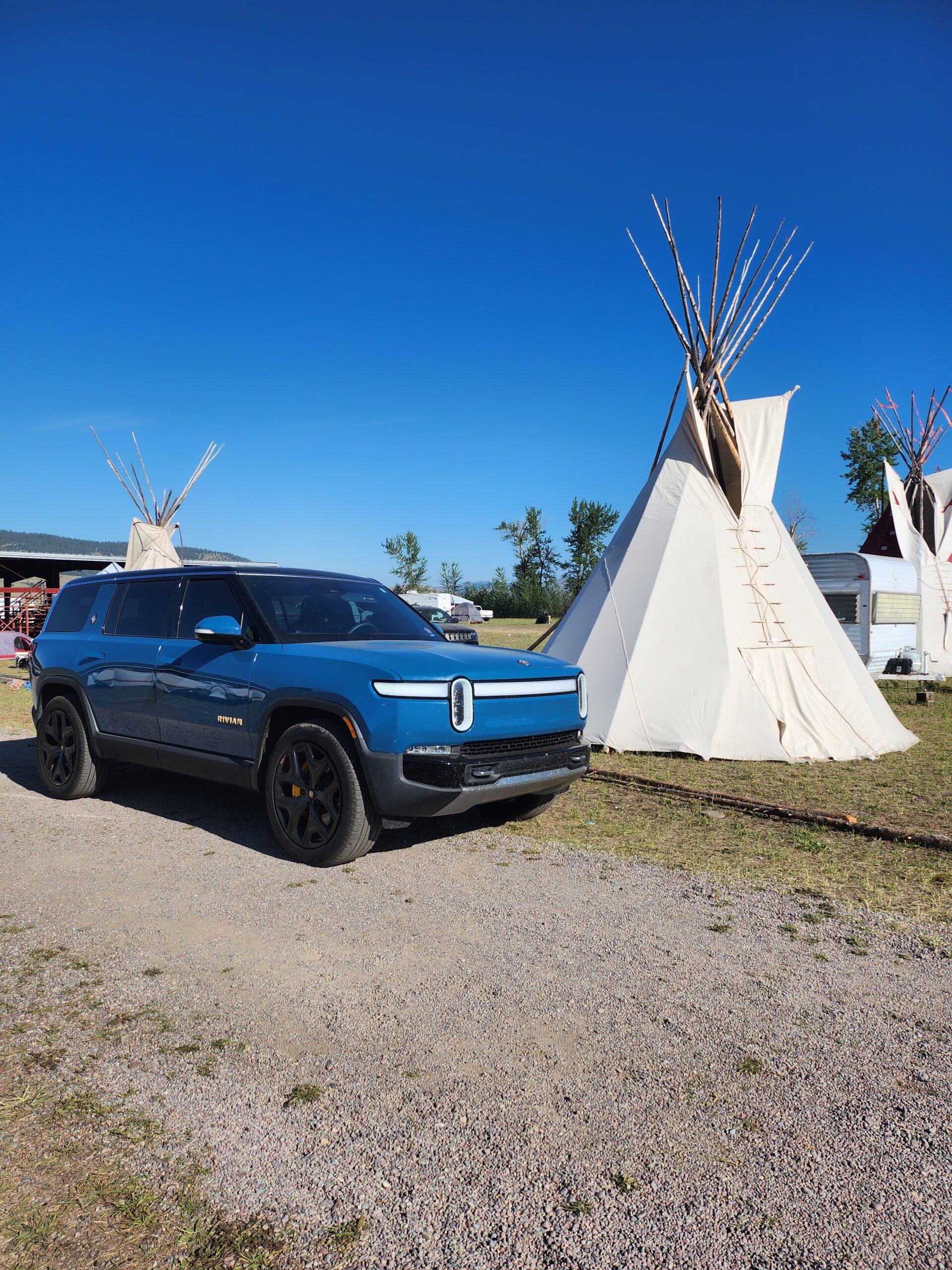A blue, Rivian electric SUV parks in front of a tipi. 