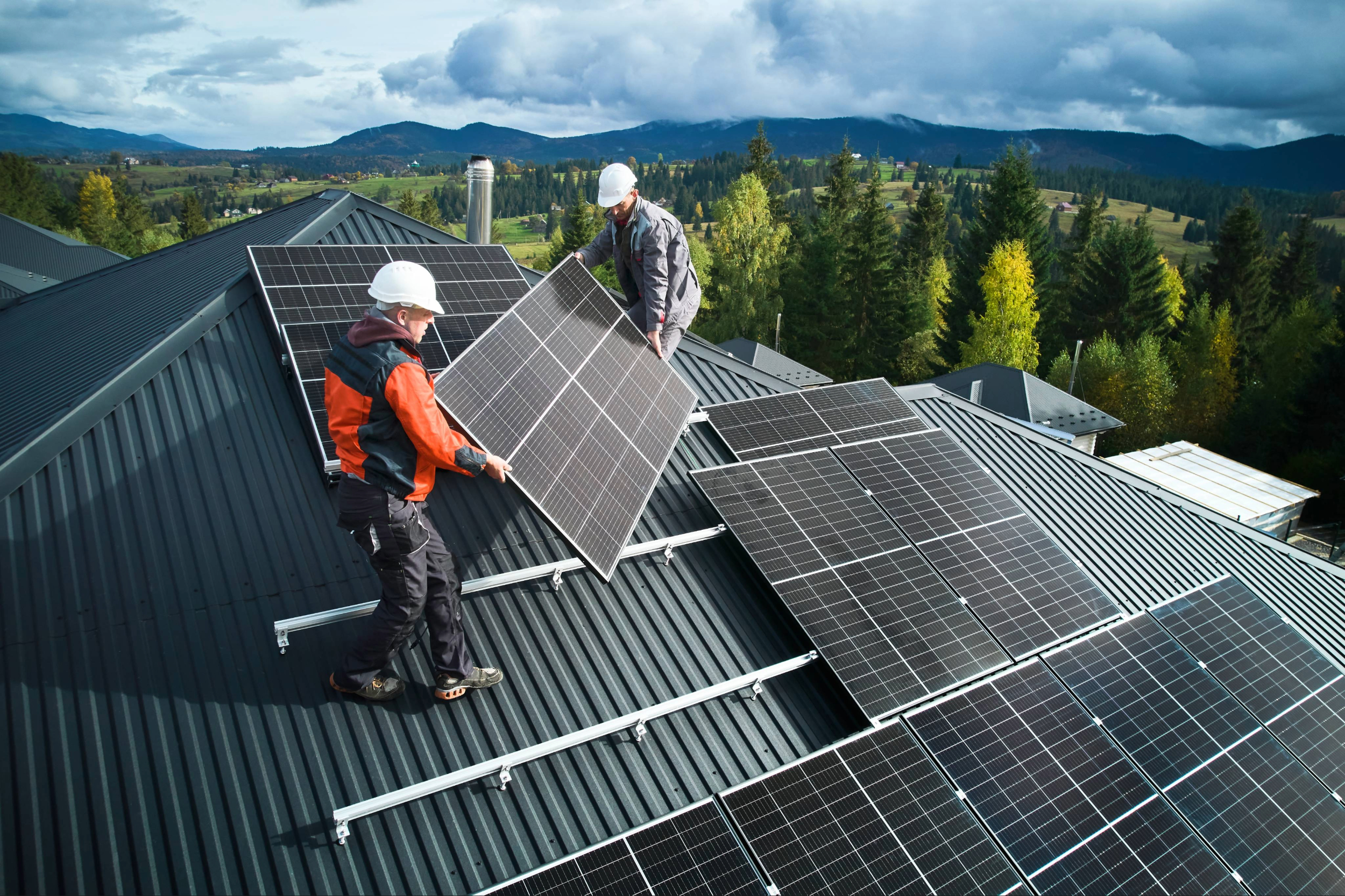 Two workers install solar panels on the roof of a building. Forests and mountains are in the background.