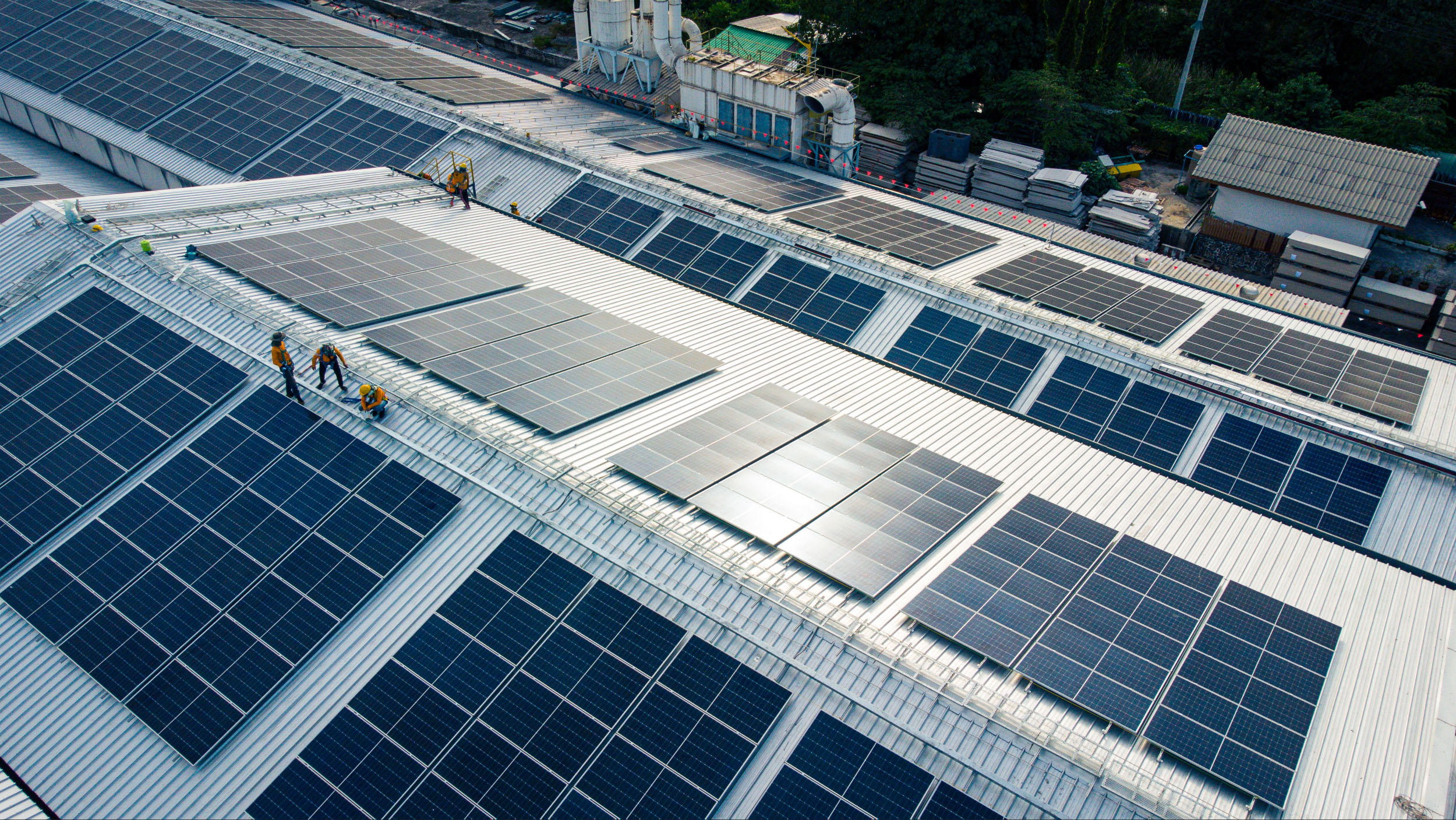 An industrial building lined with solar panels. Two workers repair a solar panel