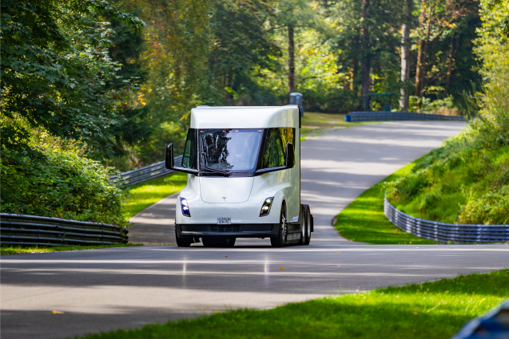 A Tesla electric truck drives on a paved road through a thick, evergreen forest.