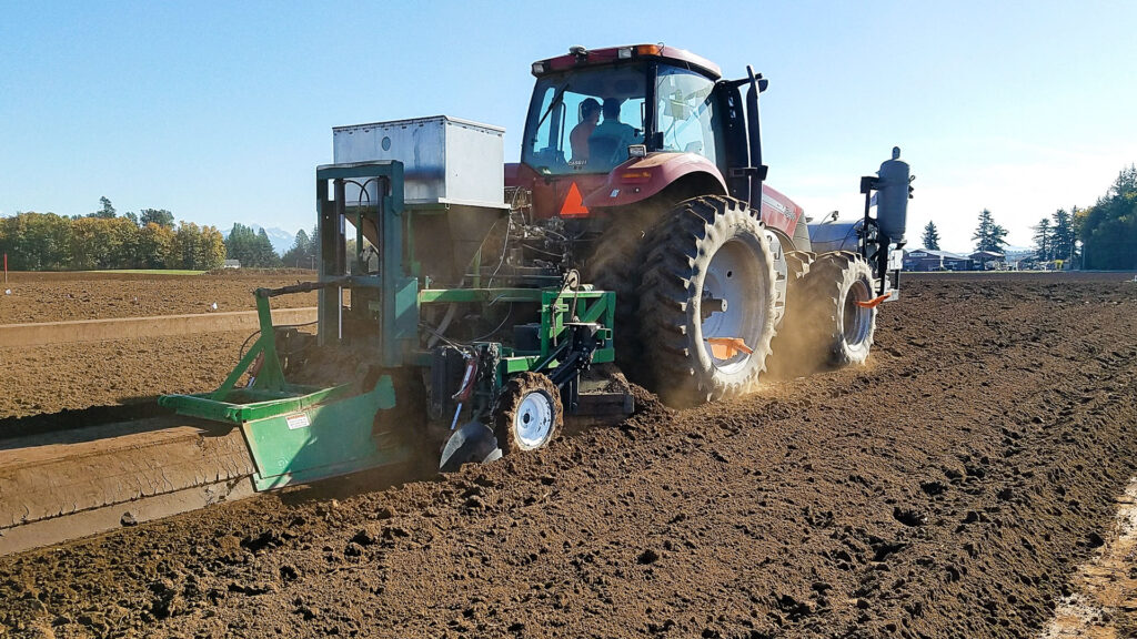 A large tractor in a field pulling a towed attachment that shapes a raised row and applies fumigant to the soil.