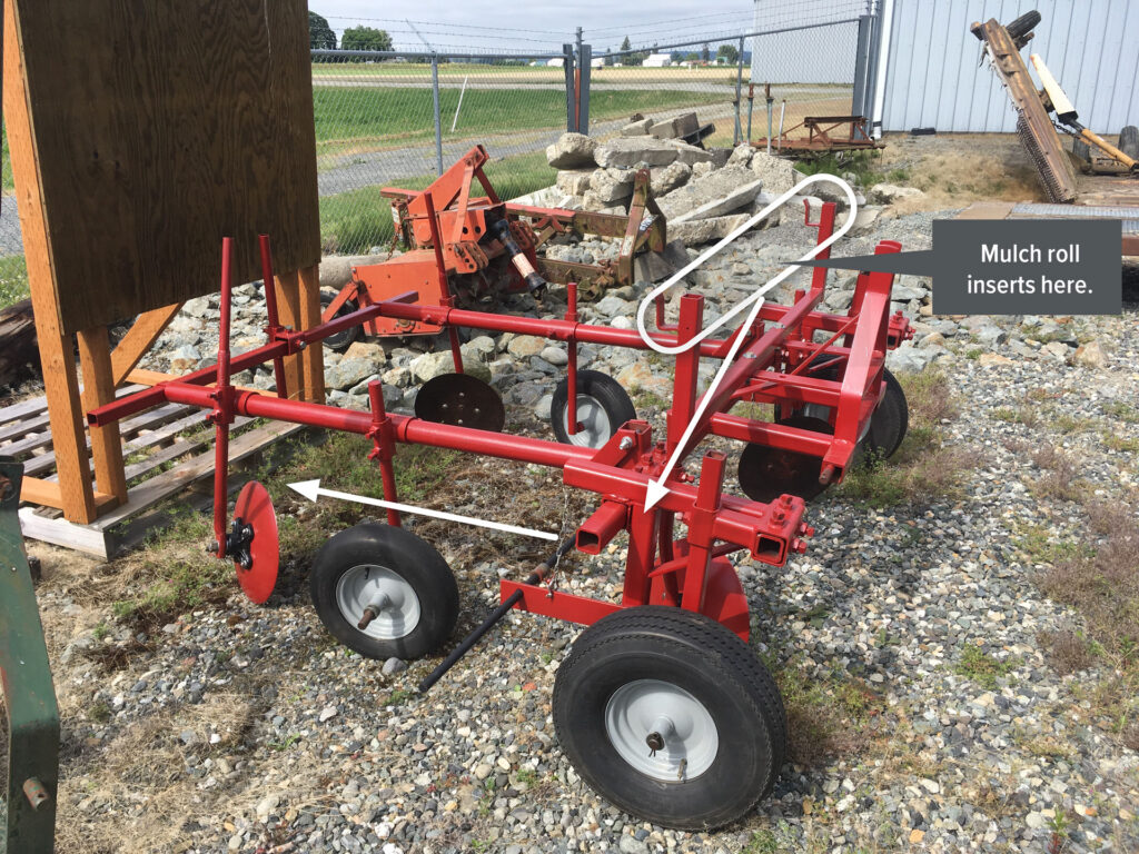 Piece of farm machinery constructed of red steel tubing. Mulch roll location indicated. White arrows indicate feed path.