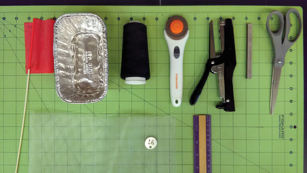 Items arranged on a self-healing cutting mat: soil flag, aluminum foil pan, thread, rotary cutter, stapler & staples, scissors, mesh fabric, metal tag #97, ruler.