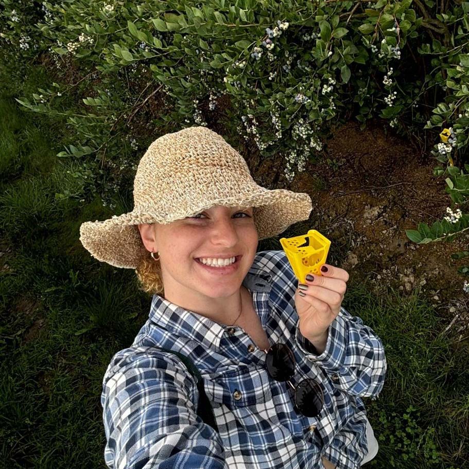 Claire, looking fabulous in a straw hat and plaid shirt is against a green leafy background holding a yellow decoy dispenser