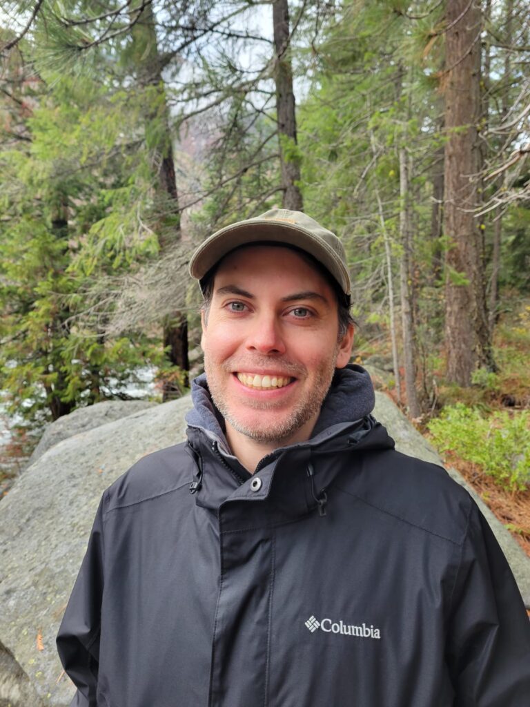 a smiling person wearing a tan ball cap and blue rain jacket standing in front of a boulder with trees in the background.