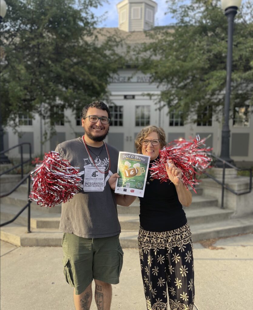 Two people stand together holding pom poms and a flier for the Beef and Brews event. Behind them is a building and trees.