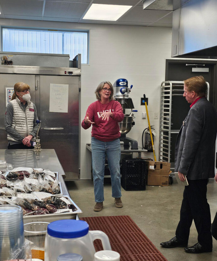 Three people stand in a room. On a metal table is food packaged in plastic ziploc bags. There is a metal cart, some plastic bins, and a large mixer in the background, as well as a large steel refrigerator. In the foreground is the top of a plastic jug as well as some plastic cups. Behind the refrigerator is a window at the top of the wall. Flourescent lights are on the ceiling.