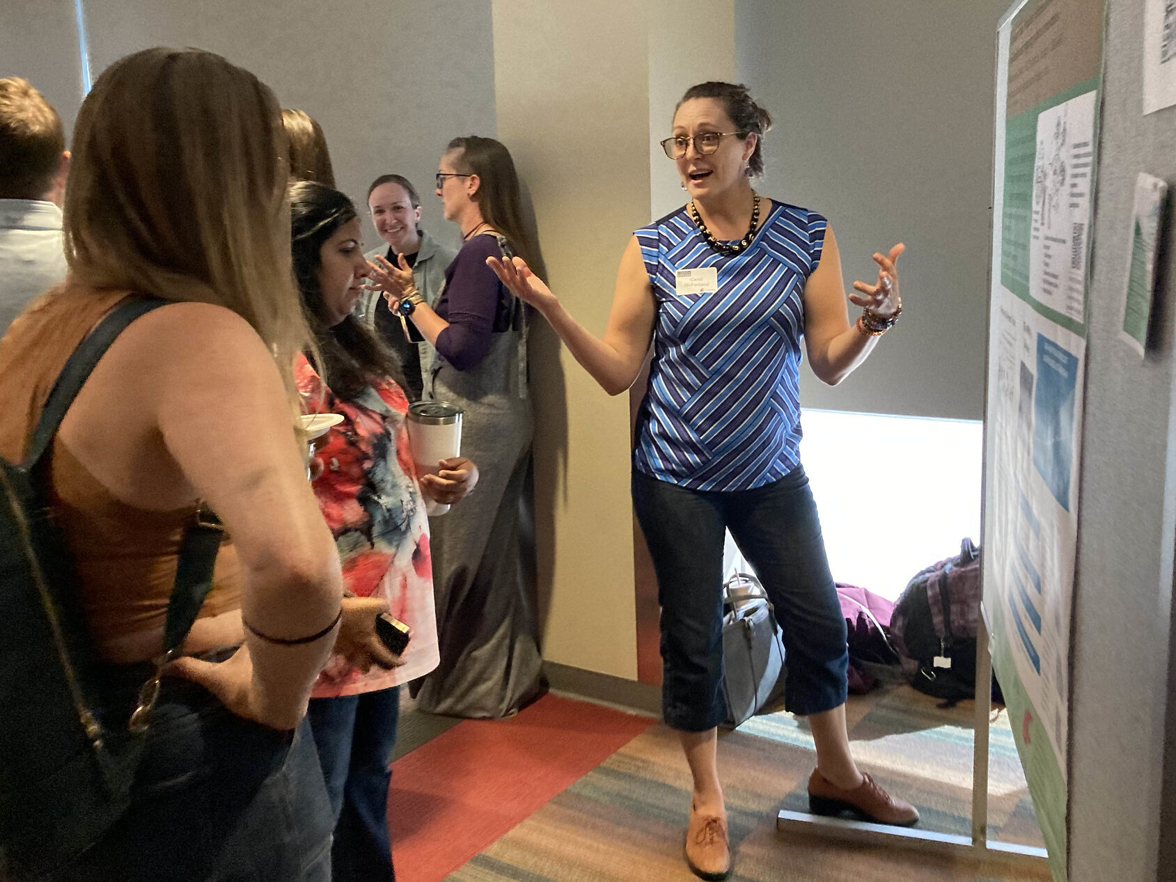 A person gestures next to a poster on a board, while a few people stand and listen. Other people stand in the background.