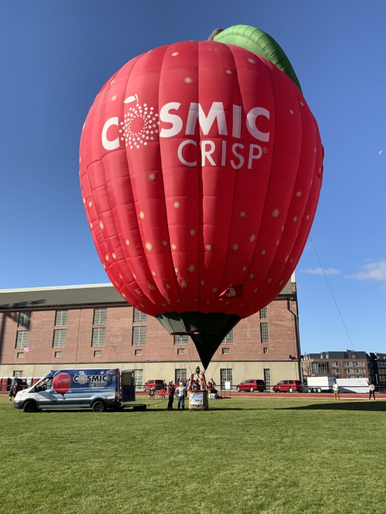 The Cosmic Crisp® balloon in a field in front of a brick building. A few people stand near the basket of the balloon, and a Cosmic Crisp® van is parked nearby. Further in the background is another brick building and a few other parked vans.