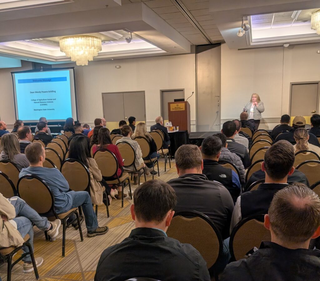 Dean Powers stands at the front of a room of a few dozen seated conference attendees. There is a slide projector screen and a podium to her right.