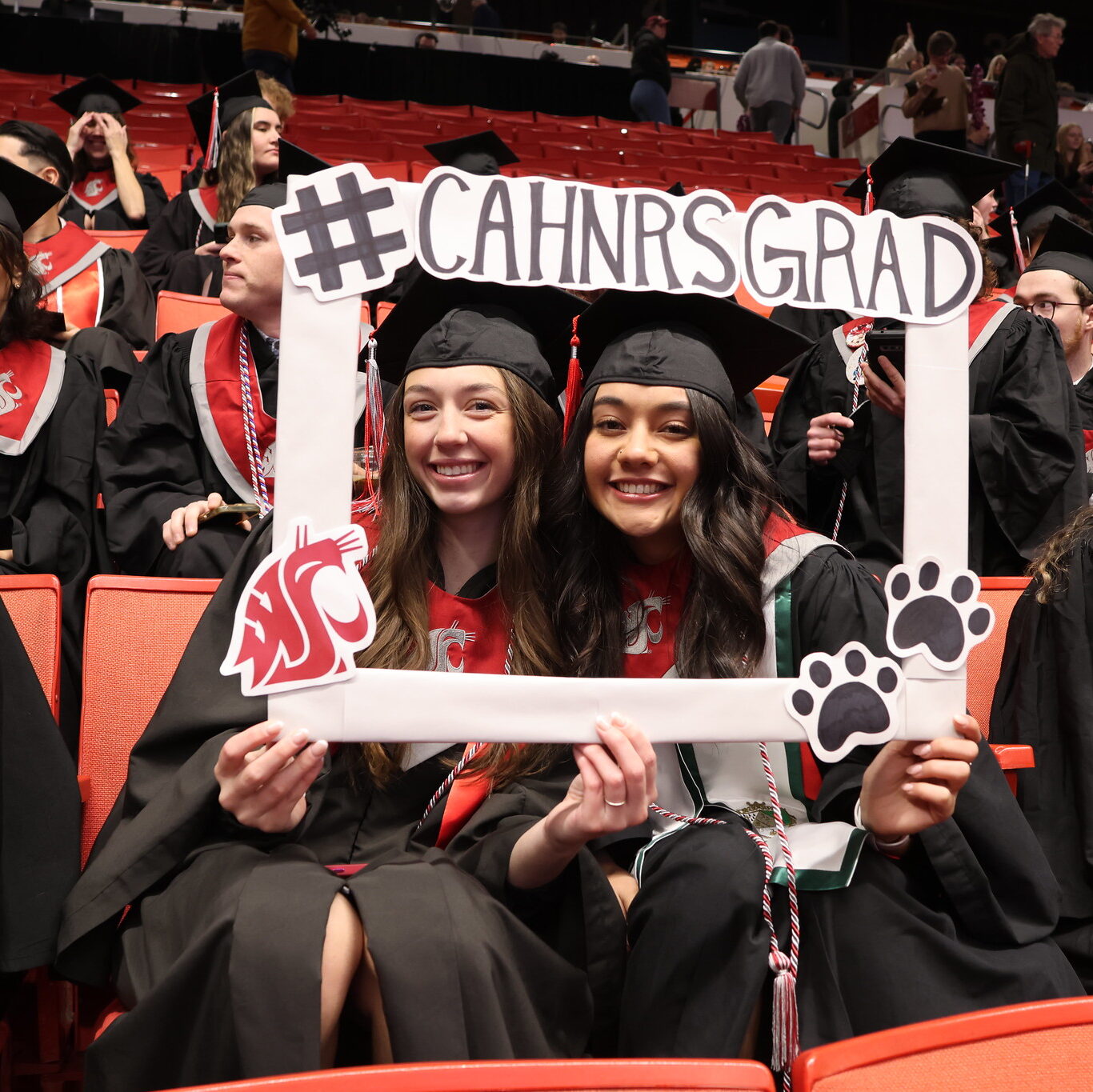 Two CAHNRS graduates in caps and gowns smile while holding a frame that says #CAHNRSGRAD with the WSU cougar head logo and two paw prints. 
