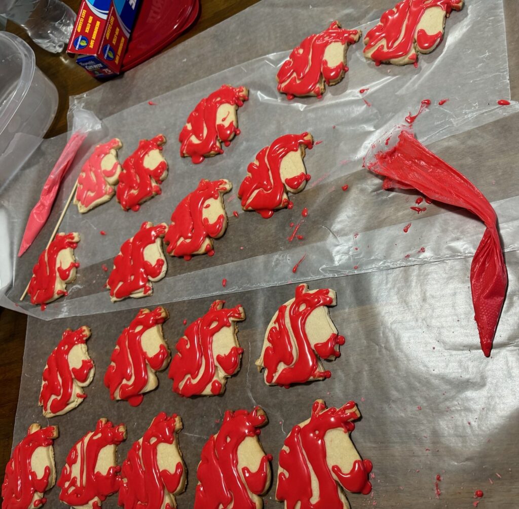 18 cookies frosted with the red WSU cougar head logo are sitting on wax paper. A frosting decorating bag is nearby.