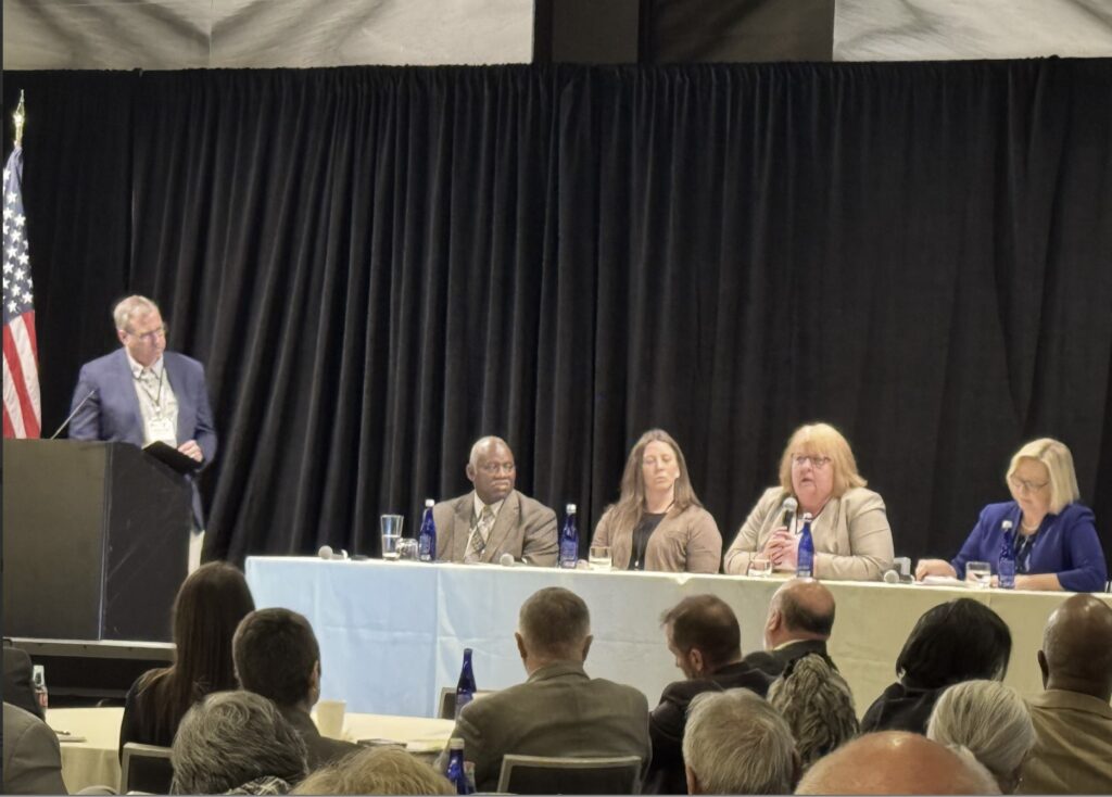 The back of a crowd who sits, listening to a panel of four people seated at a table. Another person stands at a podium and looks toward those sitting at the table.