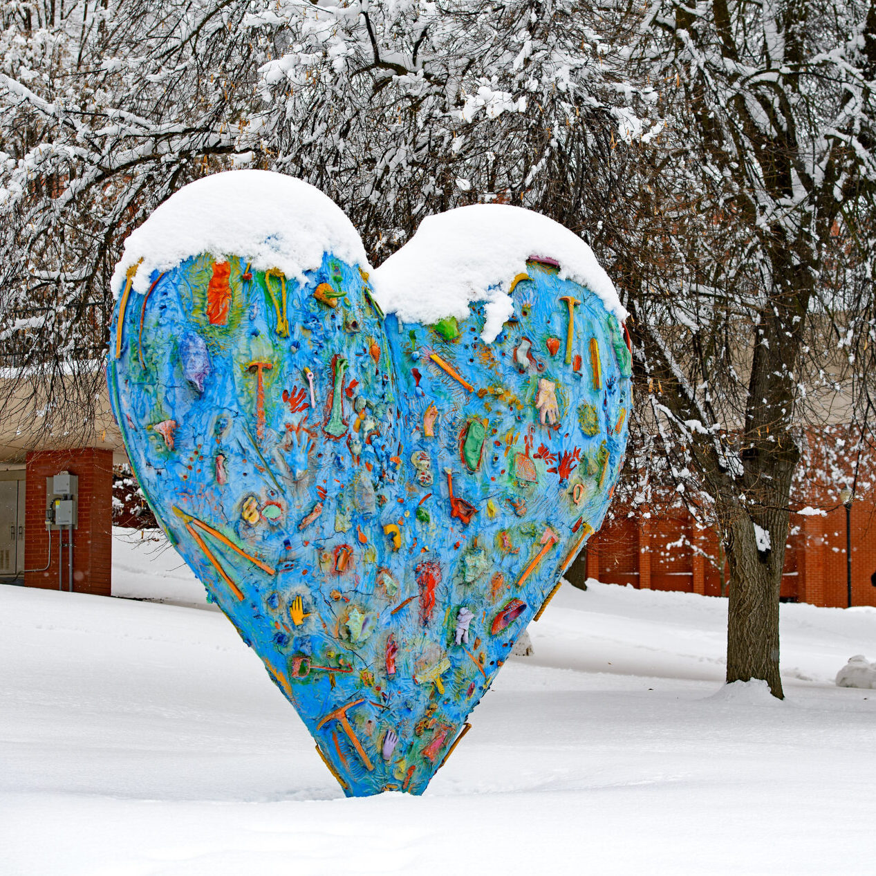 A large heart sculpture covered with hammers, shoes, hands, clamps, and flashlights, covered in a coat of snow. The ground is covered in snow and there are trees and a brick building in the background.